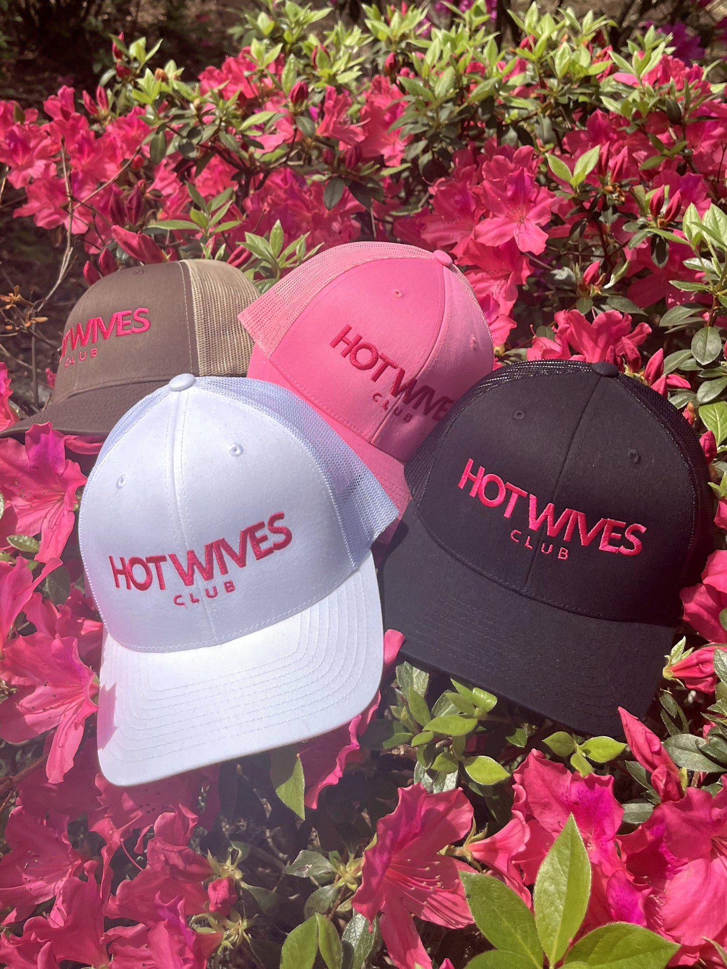 Four 'Hot Wives Club' hats in different colors on a background of pink flowers.